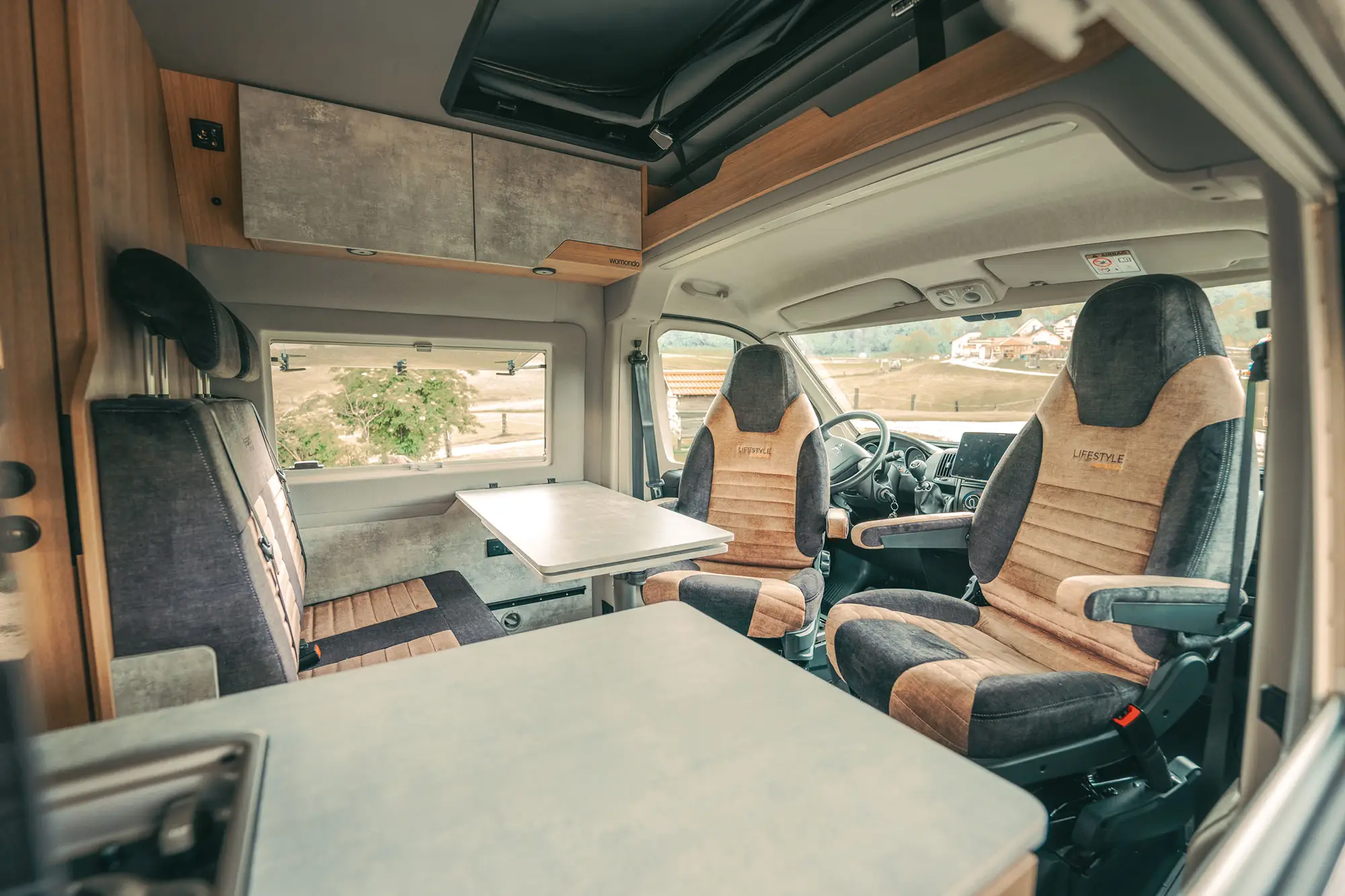 Interior of a camper van showing two brown and black driver and passenger seats labeled Lifestyle, a small table, and a bench seat by a window with outdoor views.