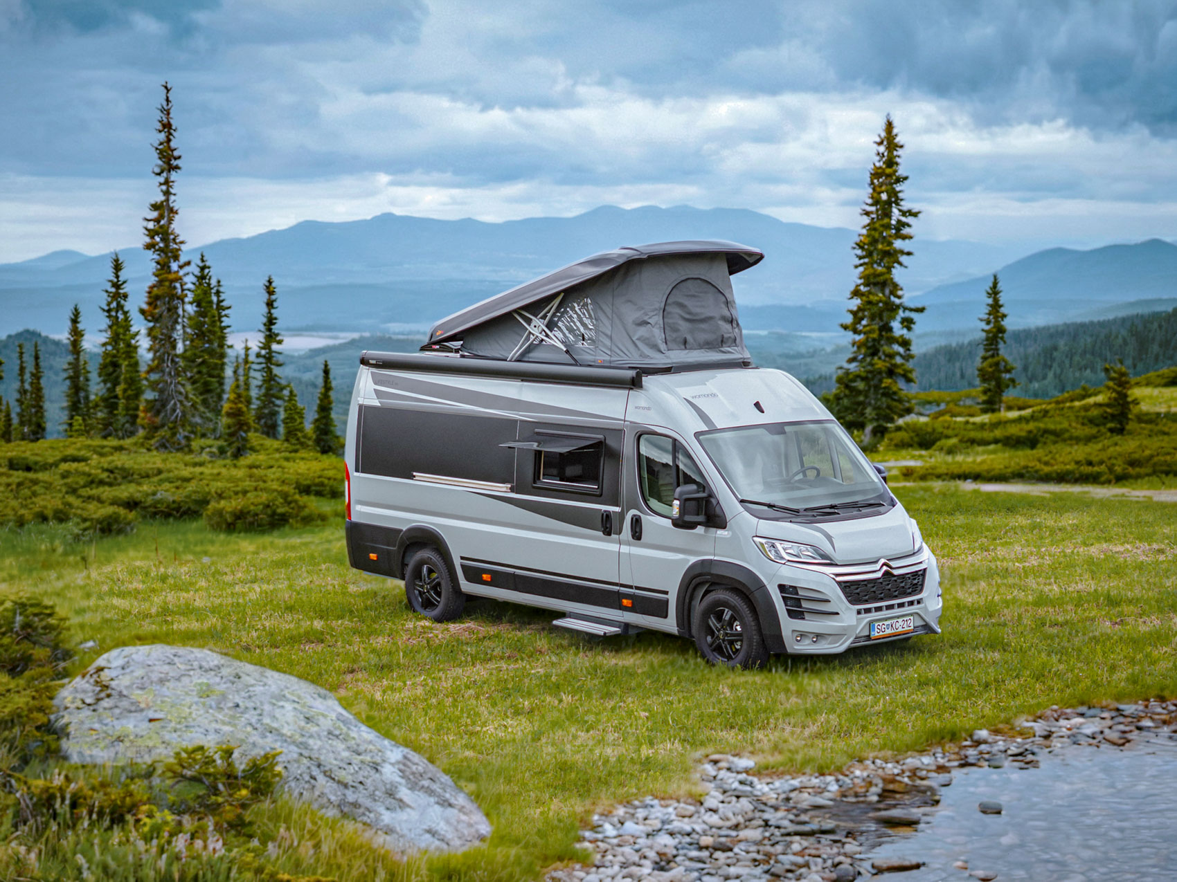 Modern gray camper van with pop-up roof parked on green grass near a small stream with pine trees and mountains in the background under a cloudy sky.