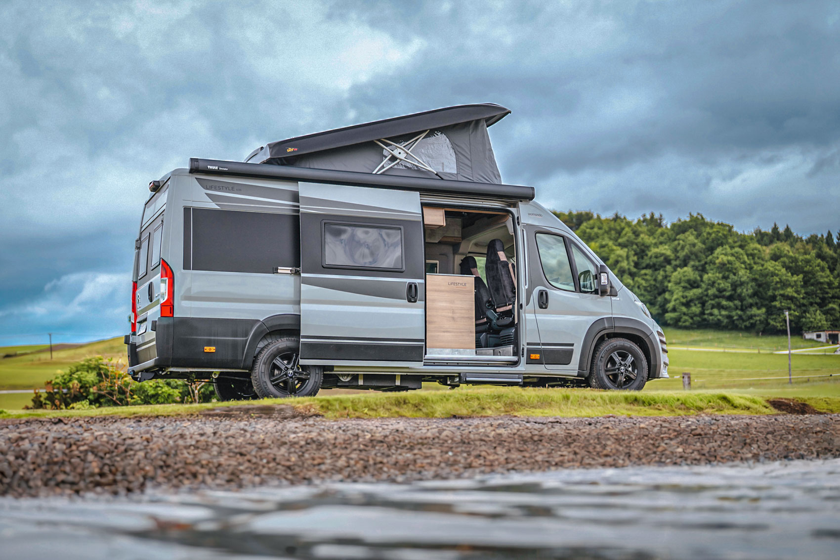 Gray camper van with pop-up roof parked by a lake with green hills and cloudy sky in the background.