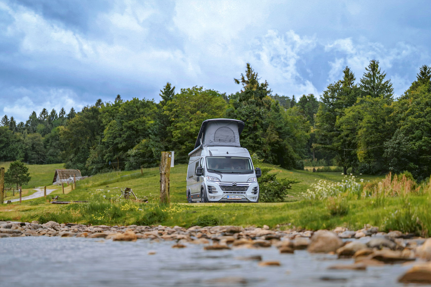 White camper van with raised roof parked on green grass near a river, surrounded by trees under a cloudy sky.
