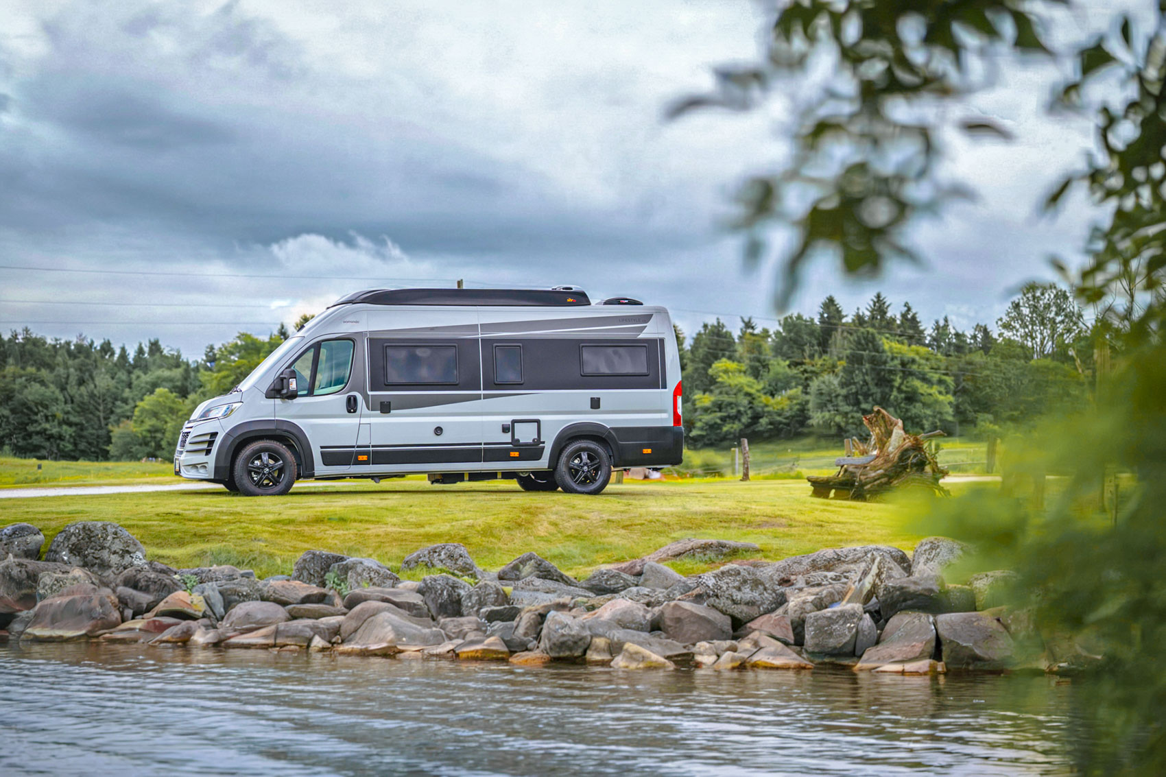 White and gray camper van parked on grassy area near a rocky riverbank with trees and cloudy sky in the background.