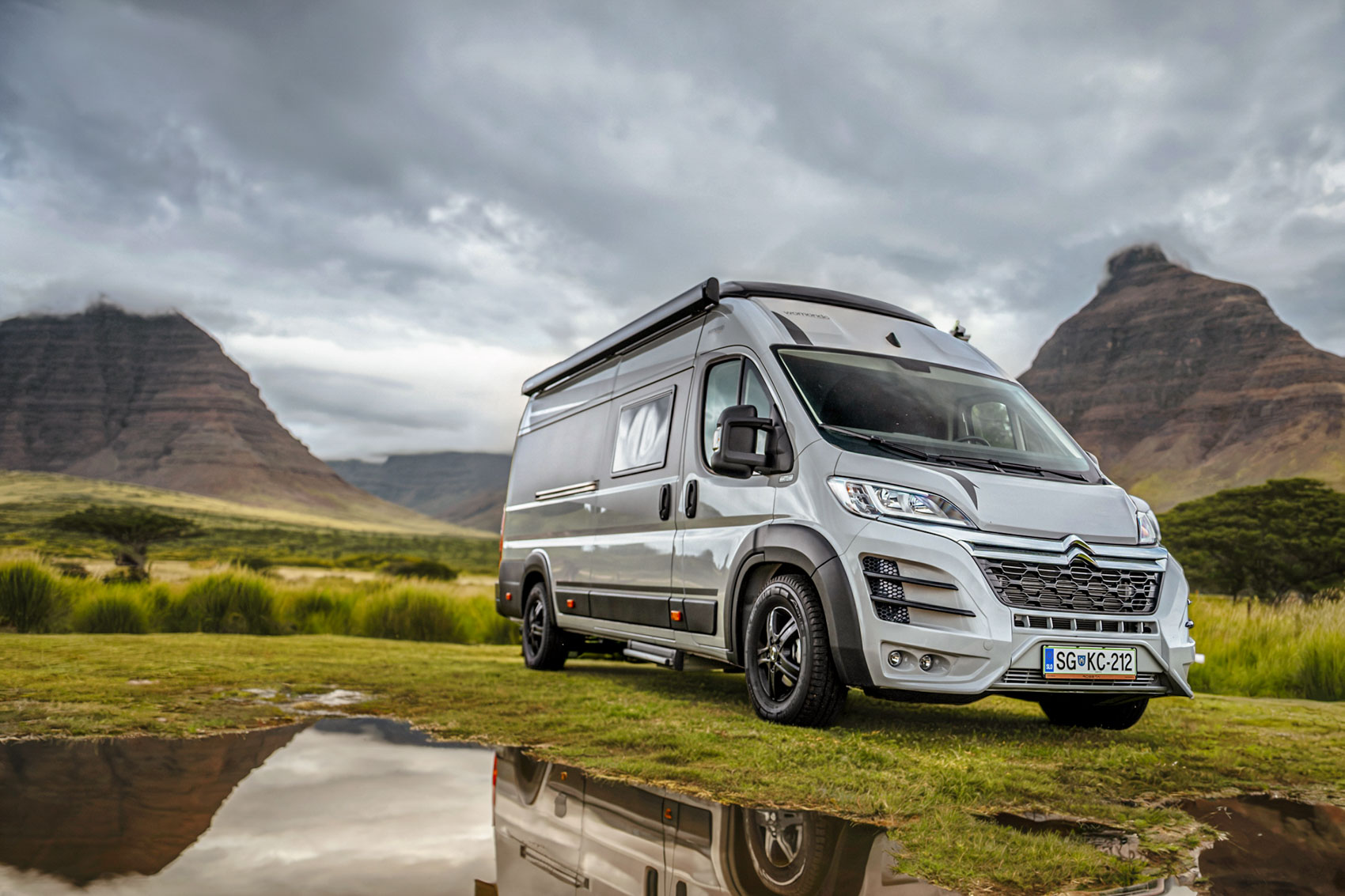 Silver camper van parked on green grass with mountains and cloudy sky in the background reflecting in a nearby puddle.